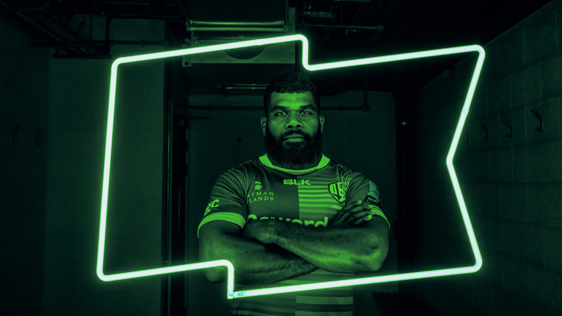 A burly bearded man in his rugby kit with arms folded front-lit by a neon green outline of a flag