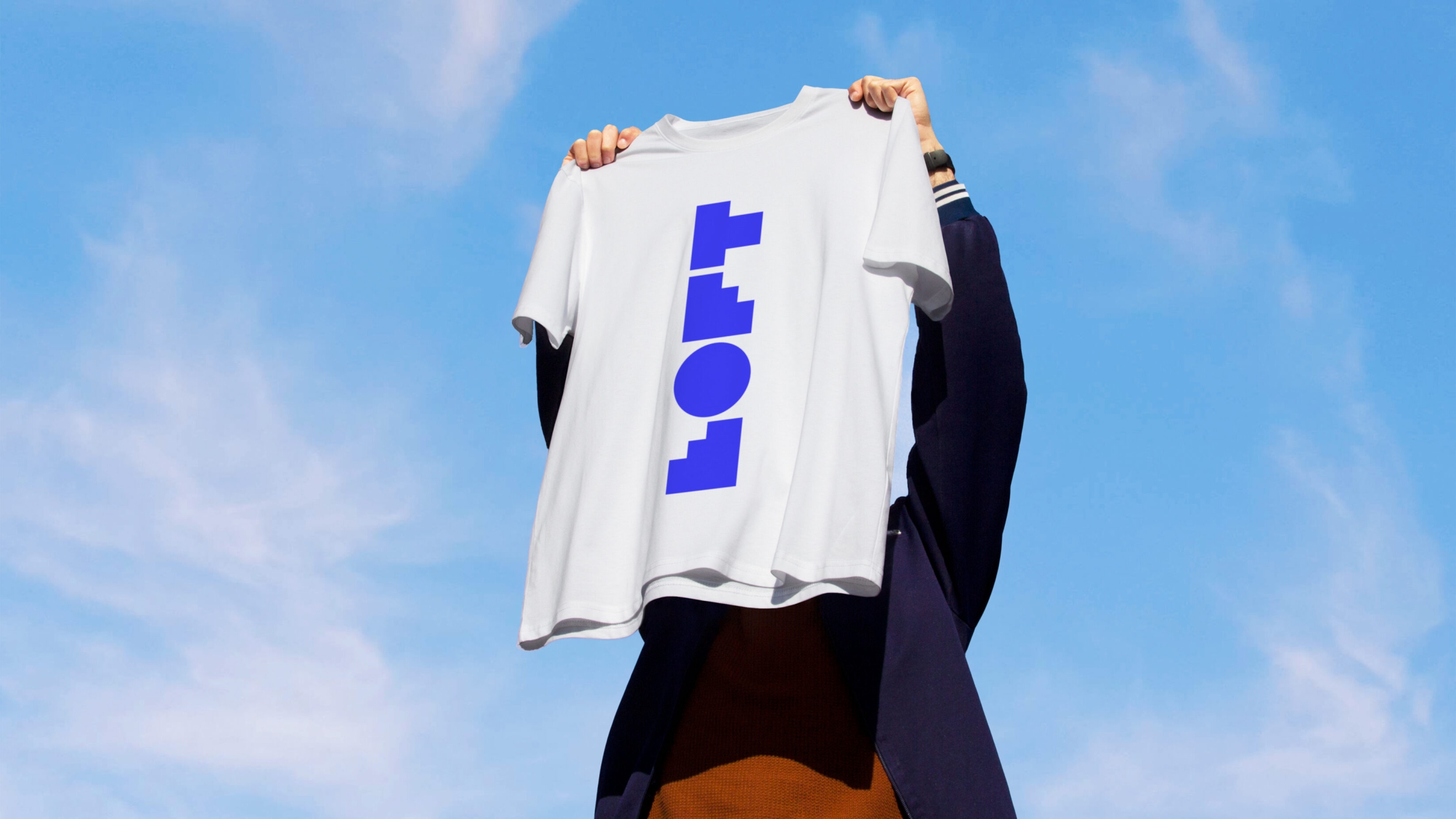 A white t-shirt with the Loft logo, now vertical, in electric deep-blue is held aloft by an individual whose face is hidden by the hanging material.