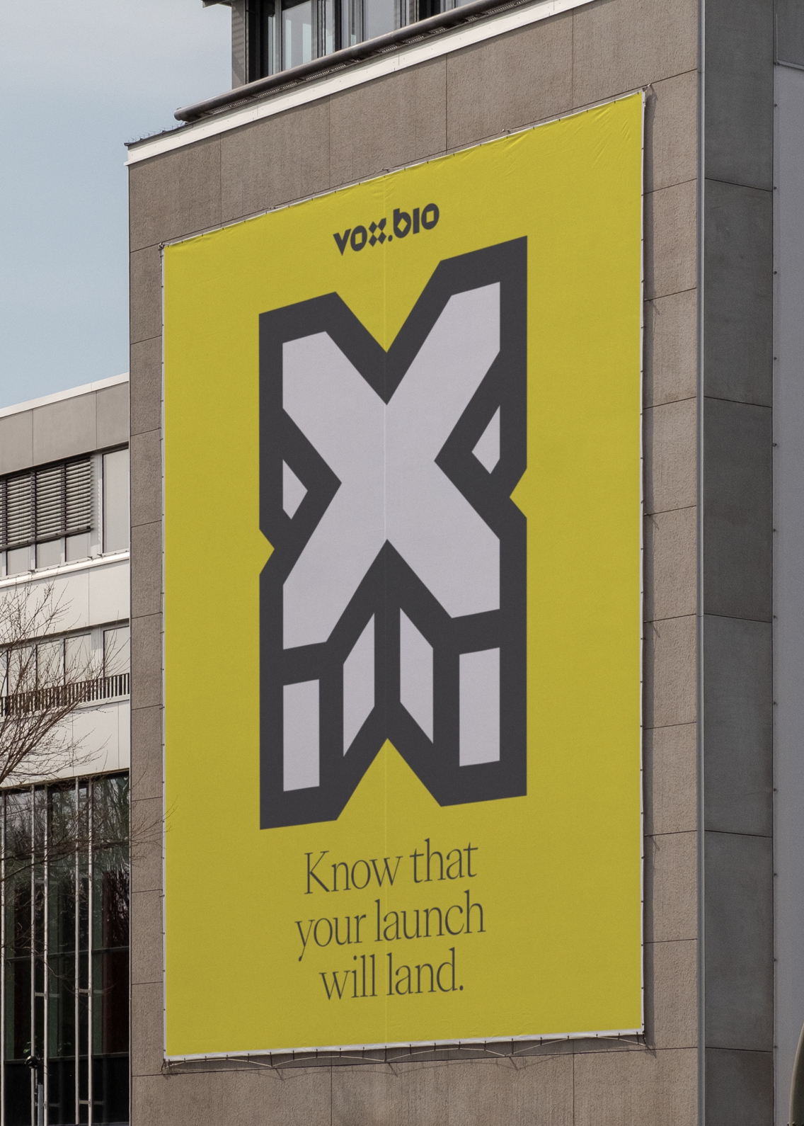 A Vox.Bio poster pasted onto the side of a concrete building. The bold white cross on yellow background with black text is a stark contrast to the industrial building it is hanging on.