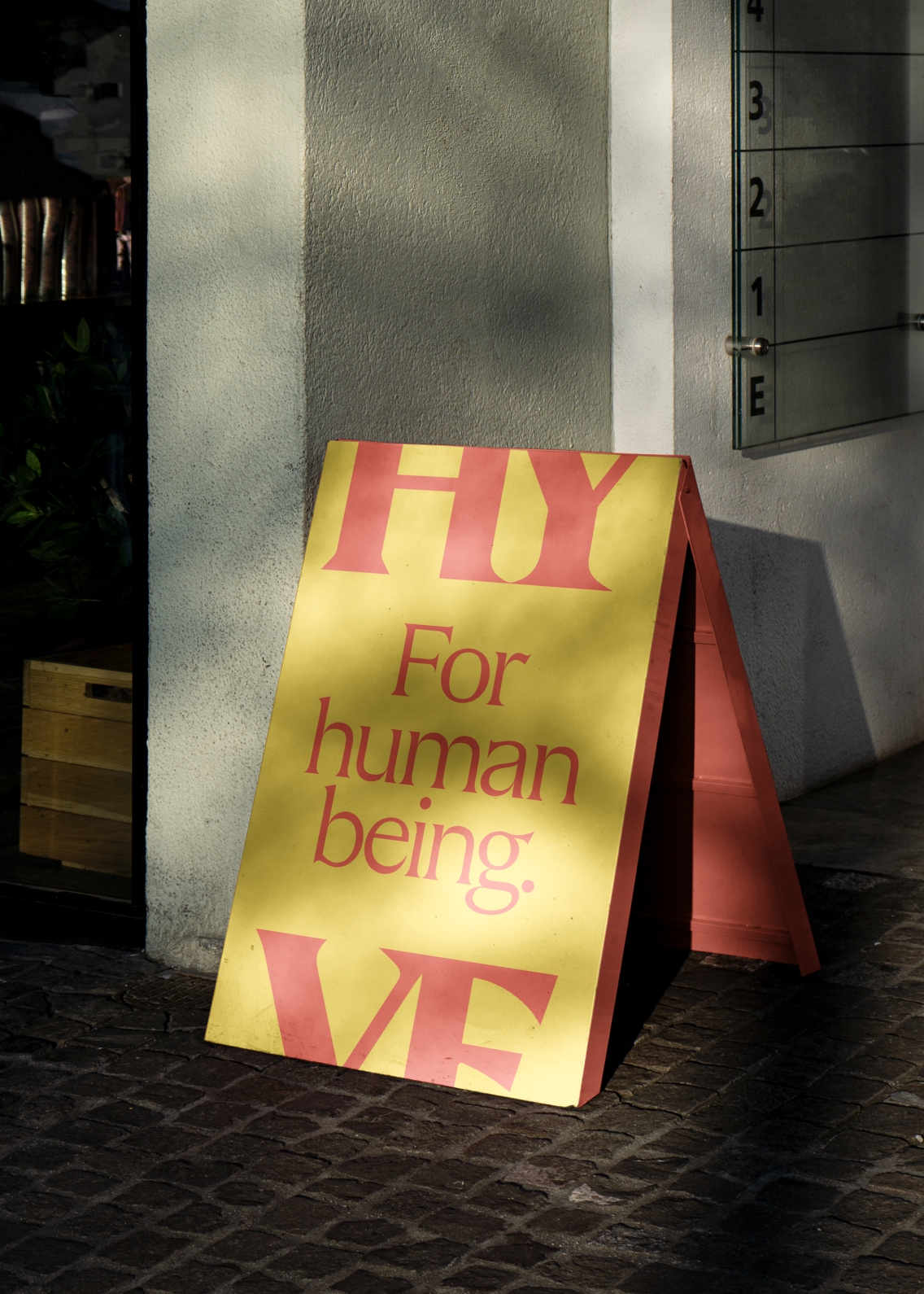 'For human being' large type a-board in red on a yellow background, framed with a HY at the top and VE at the bottom.