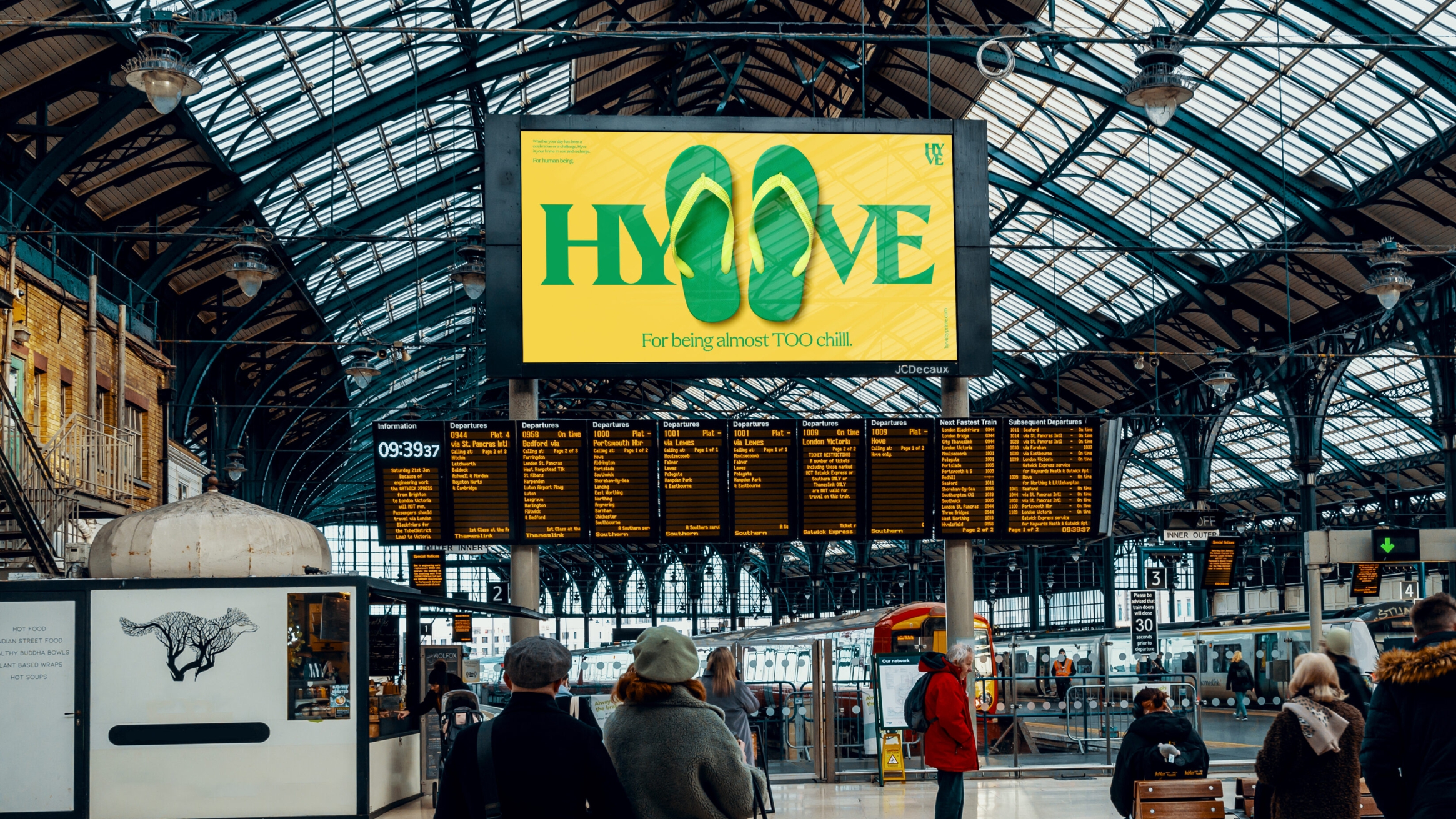 Billboard at Brighton tain station, with a large HYVE logo and a pair of green and yellow flip flops in the centre, splitting the wording.