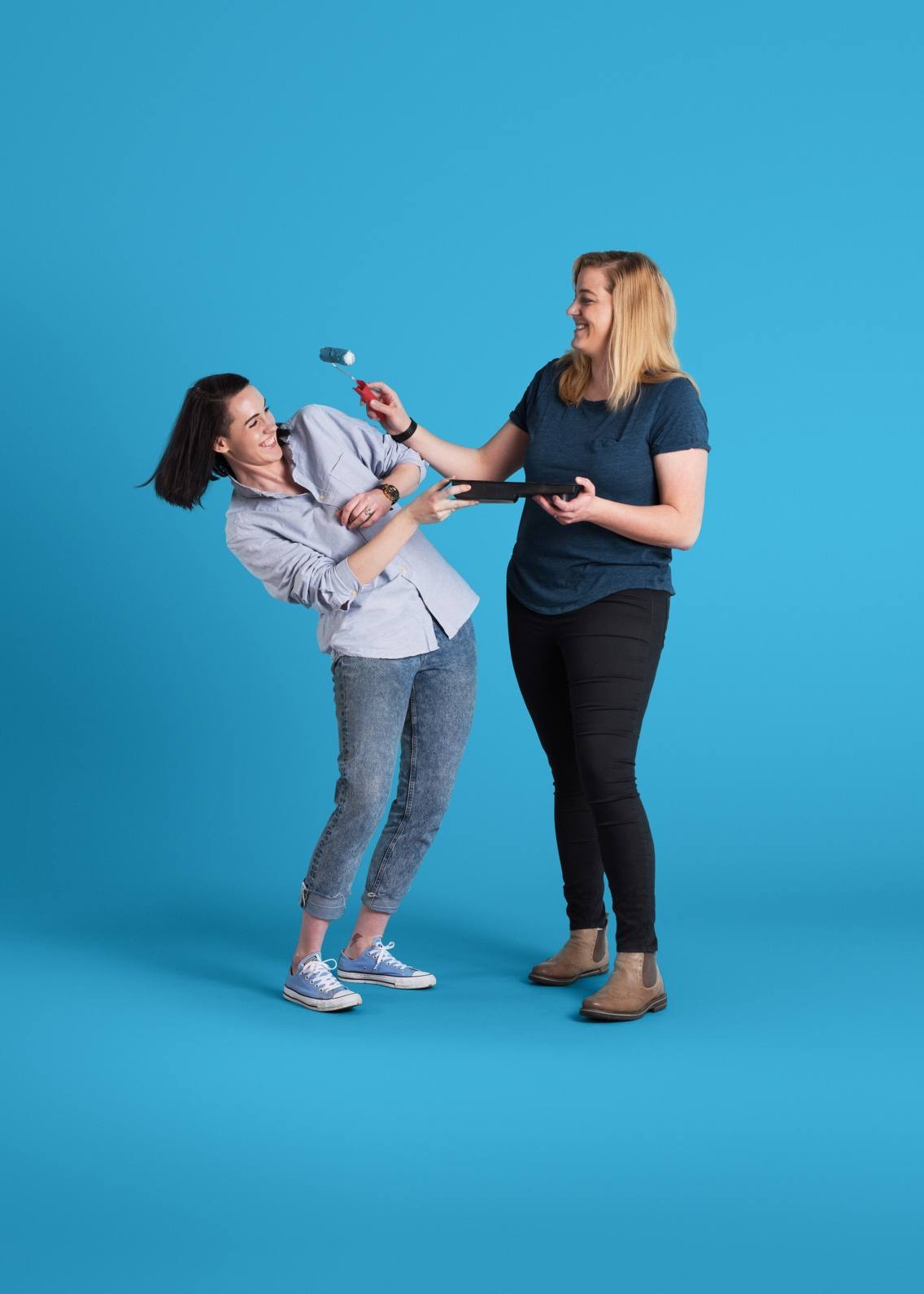 Two women in their late 20's playfully engage while holding a paint tray and roller. They have been photographed on a blue background.