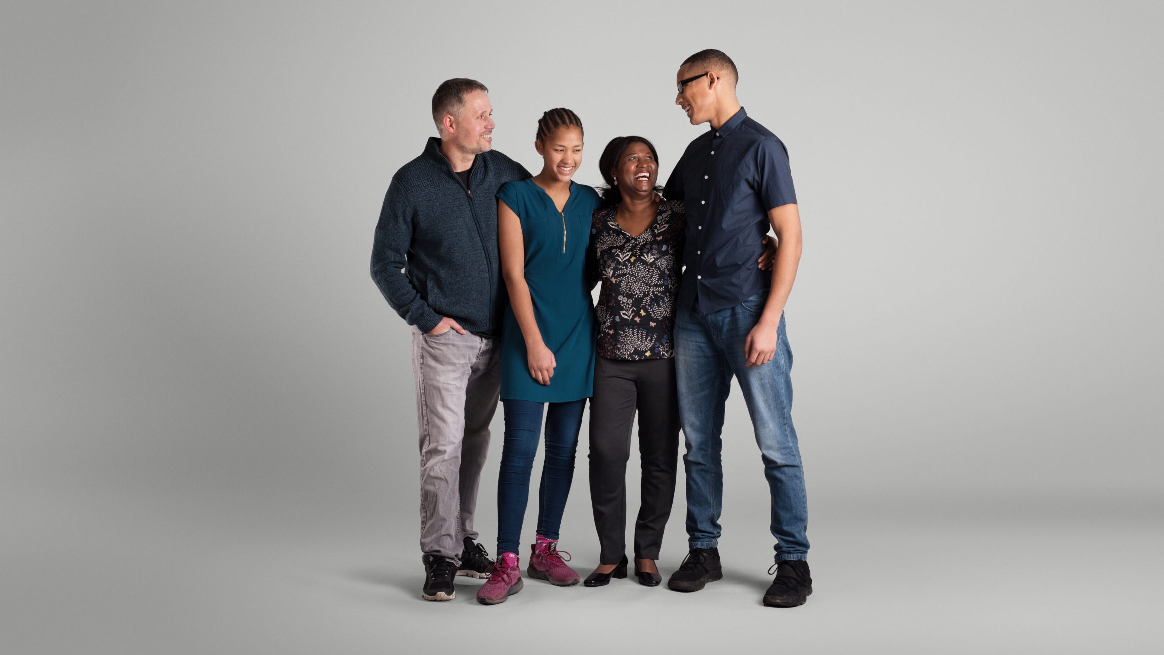 Multi-cultural family consisting of a mum, dad, son and daughter stand together while laughing. They have been photographed on a grey background.
