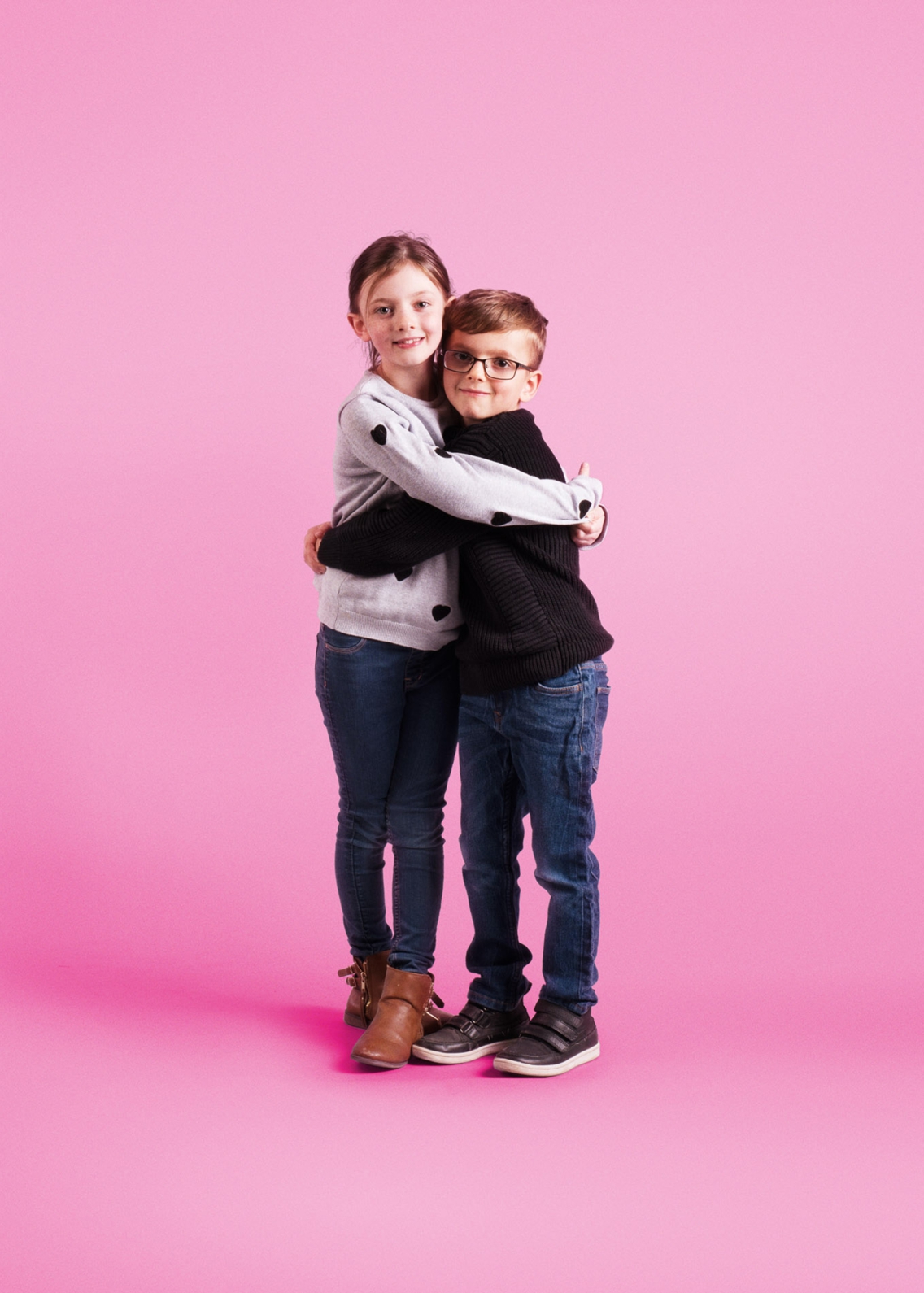 A young brother and sister hugging and smiling. They have been photographed on a pink background.