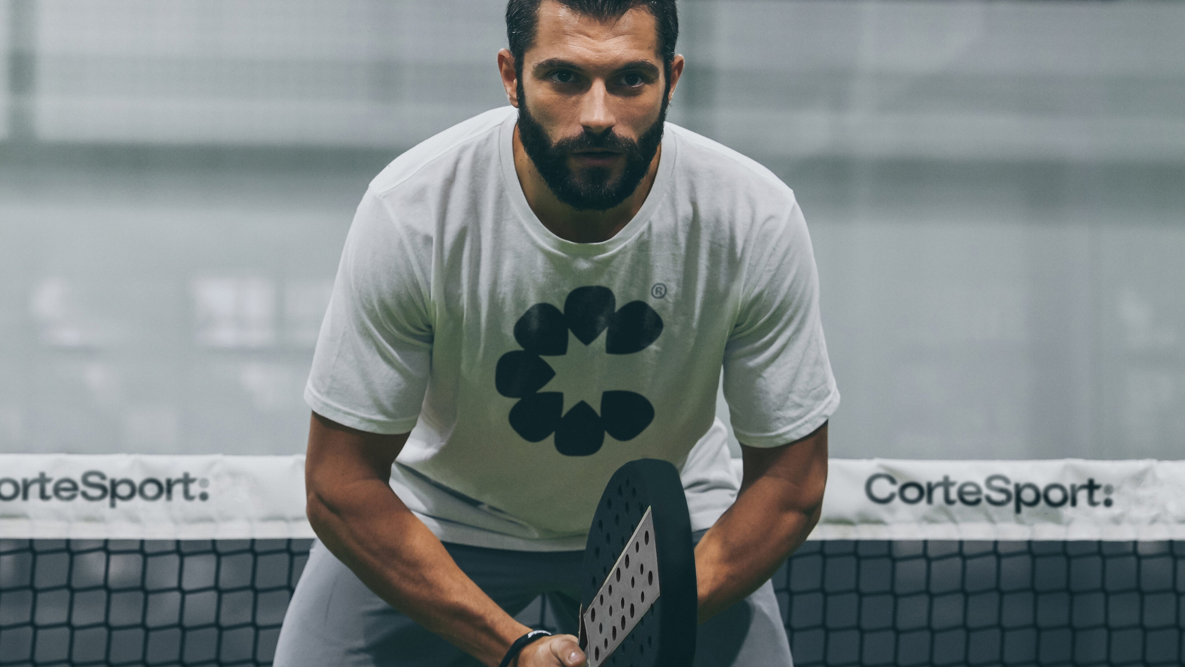 Male padel player on court, waiting to receive a serve. He is wearing a white t-shirt, with a large CorteSport icon on the front.
