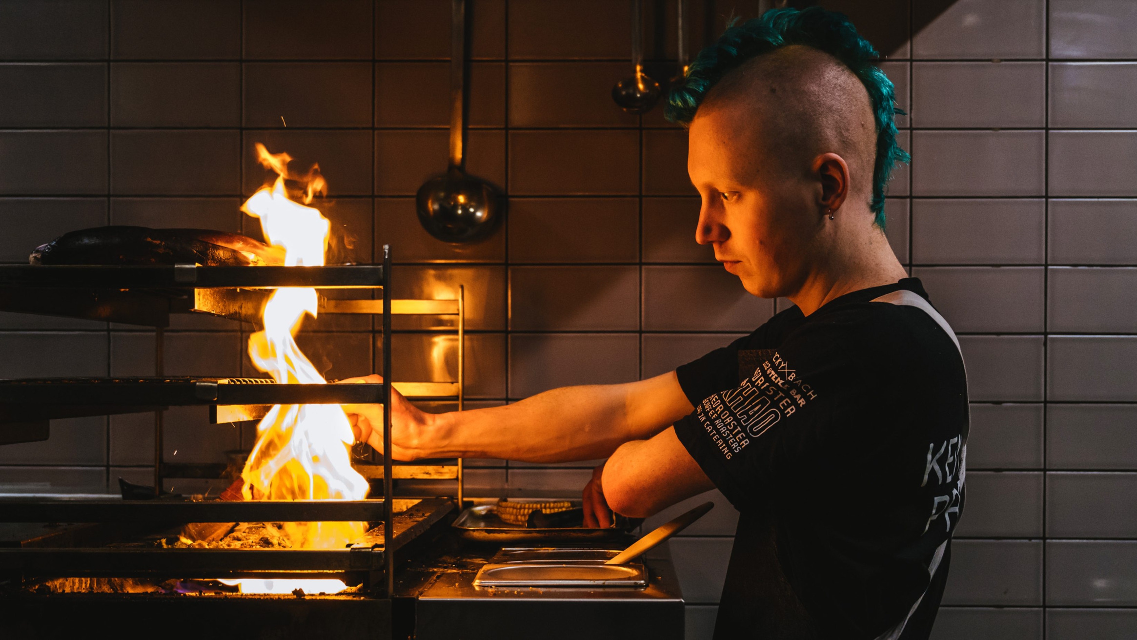 A chef with a turquoise mohawk is cooking with an open flame in the restaurant's kitchen. The light of the fire is reflecting off of his face highlighting his focus to the task at hand